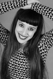 A smiling woman with long hair and bangs poses playfully, wearing a patterned black and white top. The image conveys a cheerful demeanor and artistic flair.