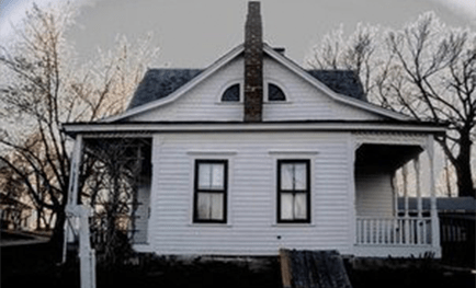 Historic white house with a central chimney, featuring large windows and a porch. Set against a backdrop of trees, it highlights early 20th-century architecture.