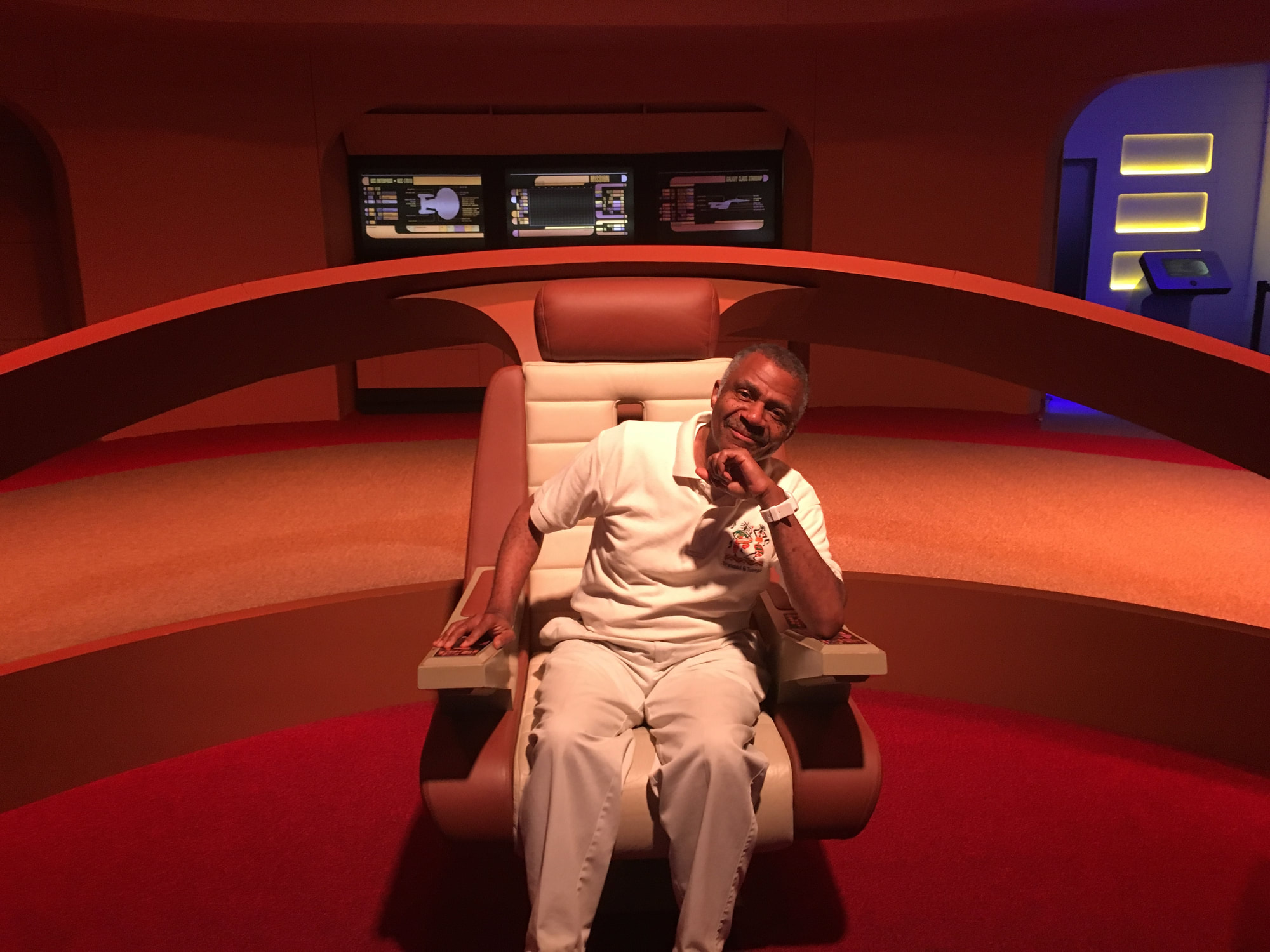 A man sits thoughtfully in a captain's chair on a sci-fi set, surrounded by high-tech displays and futuristic design elements, evoking a space exploration theme.