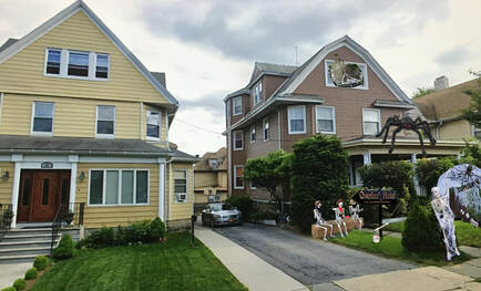 Two neighboring houses decorated for Halloween. The yellow house has a well-maintained lawn, while the brown house features spooky decorations, including skeletons and a large spider.
