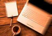 Blurred laptop, notepad, and a cup of coffee on a wooden table, suggesting a cozy workspace ideal for writing or brainstorming.